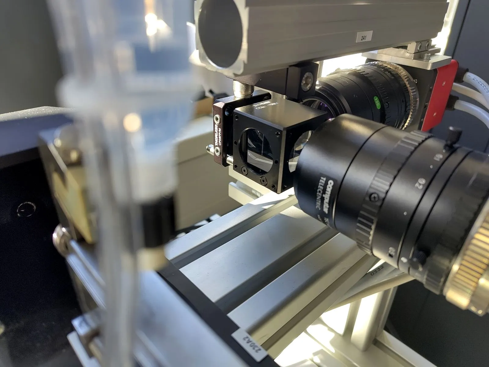 A compact HSCAM camera with a telecentric lens, aligned to a beam-splitter cube for coaxial backlighting, mounted on a precision motion-stage rig.