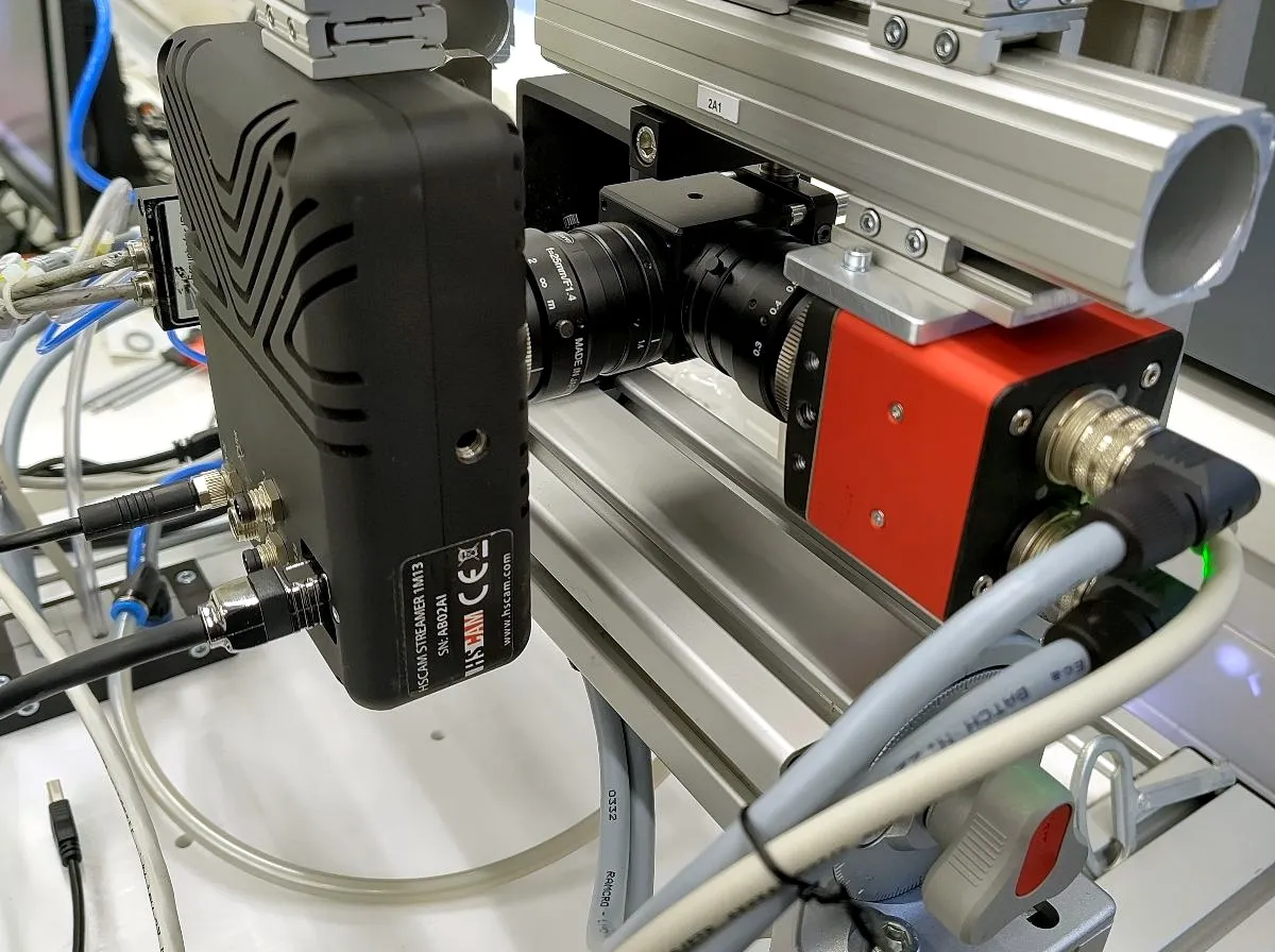 HSCAM STREAMER 1M13 camera (labelled, serial AB02A1) integrated into a precision motion-stage test rig alongside a compact camera variant, in a real lab setup.